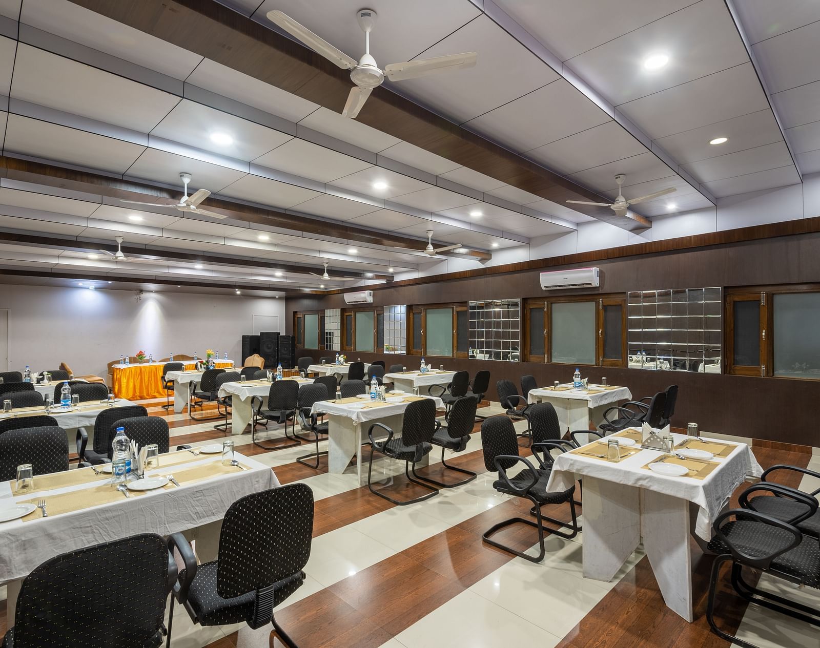 A well-lit conference room with rows of tables and chairs, featuring a projector screen and a fan - DLS Meghavan Resort